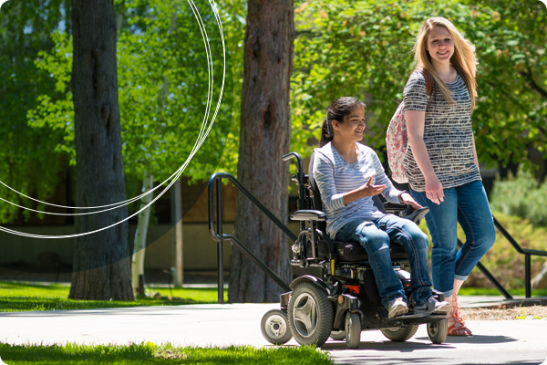 两名俄勒冈州中部社区的学生在人行道上走动和交谈，一名学生坐在电动轮椅上